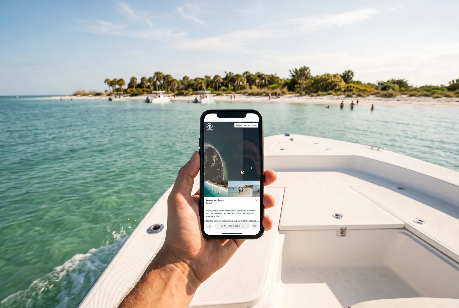 A hand holding a phone at the bow of a boat, showing the Nebo Discovery card for Egmont Key Beach, with that palm-fringed beach and anchored boats just ahead