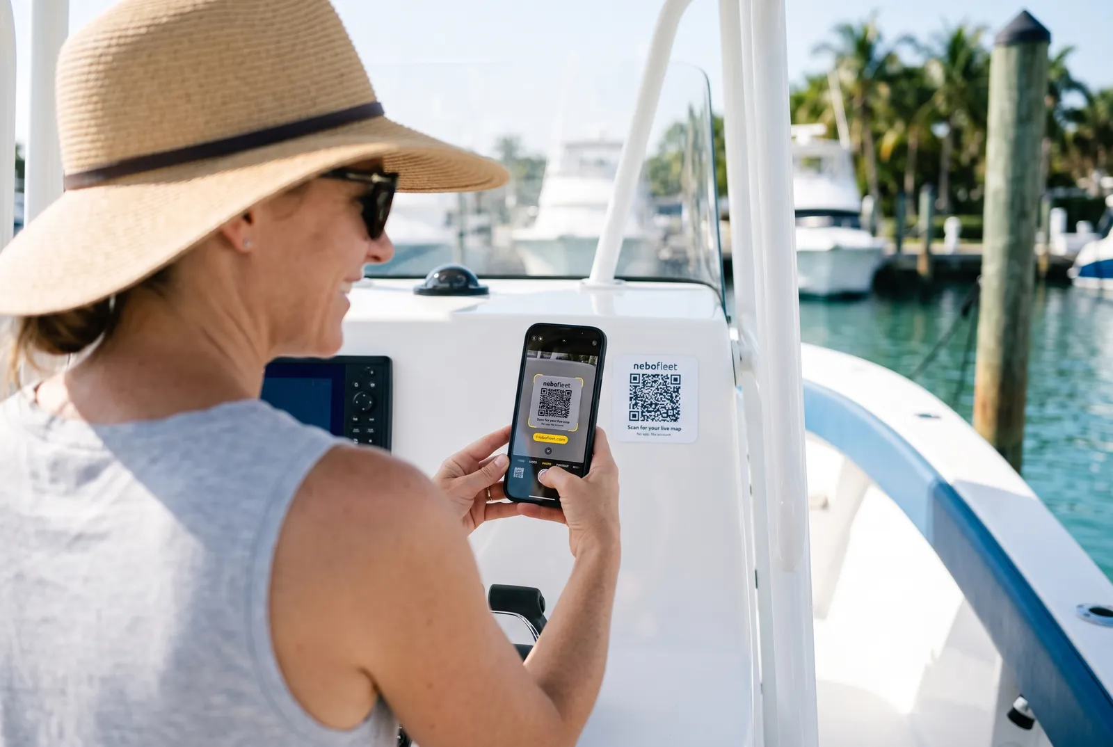 A woman in a sun hat at the helm of a center-console boat, using her phone's camera to scan the NeboFleet QR code on the console